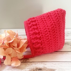 May include: A bright hot pink crocheted bonnet featuring a textured stitch pattern and long matching ties. The bonnet is displayed alongside a cluster of soft peach-colored artificial hydrangea flowers, highlighting the vibrant yarn color.