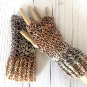 May include: Crocheted fingerless gloves in shades of brown, tan, and gray. The gloves have an openwork design and are displayed on a mannequin hand. The wrist cuffs have a ribbed texture.