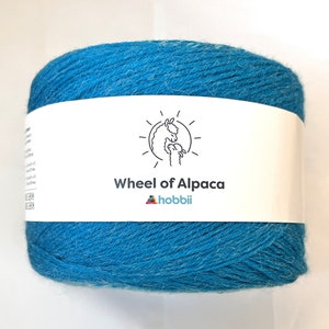 May include: A ball of turquoise yarn with a white label wrapped around it. The label features a line drawing of two alpacas and the text "Wheel of Alpaca" and "hobbii". The yarn is a vibrant blue color.