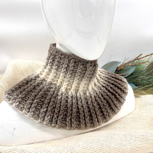 May include: A crocheted cowl in brown and cream ombre yarn. The cowl is displayed on a white mannequin.