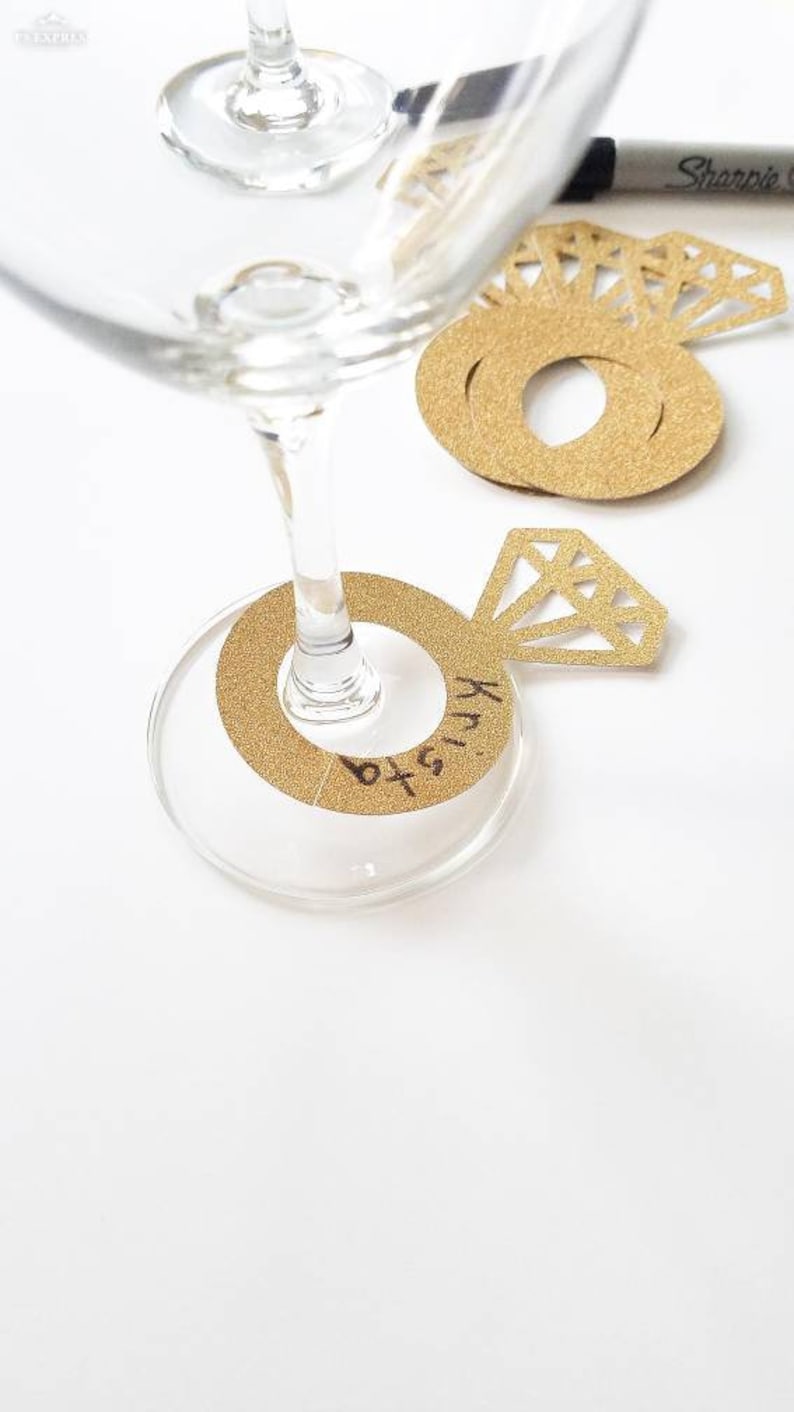 May include: Gold glitter ring-shaped wine glass charms with a diamond-shaped accent. One charm is attached to a wine glass, with the name "Krista" written on it. A Sharpie pen is visible in the background. Perfect for a wedding or engagement party.