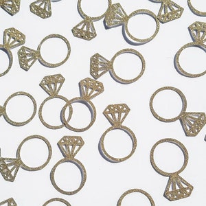 May include: Gold glitter engagement ring confetti. The confetti is in the shape of a ring with a diamond. The confetti is scattered on a white background.