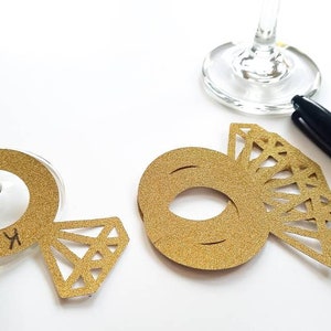 May include: Gold glitter ring-shaped place cards with a diamond design, used for weddings or events. One card has the name "Krista" written on it. A black Sharpie pen is also present.