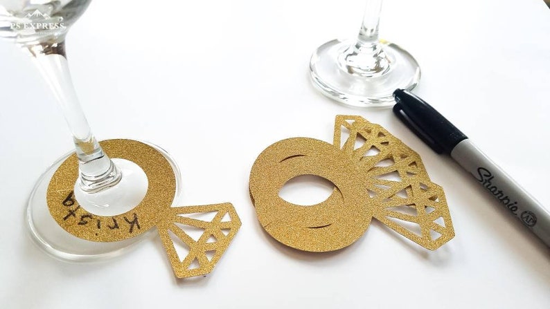May include: Gold glitter ring-shaped place cards with a diamond design, used for weddings or events. One card has the name "Krista" written on it. A black Sharpie pen is also present.