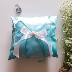May include: A light blue satin pillow with a white satin bow and a pearl accent. The pillow is trimmed with a row of white pearls.