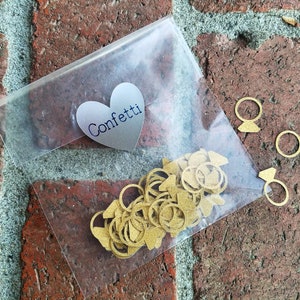 May include: A clear plastic bag filled with gold confetti shaped like rings. The bag has a silver heart-shaped sticker with the word "Confetti" printed on it.