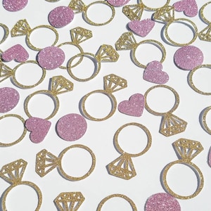 May include: Gold and pink glitter confetti featuring ring and heart shapes. The confetti is perfect for a wedding, engagement, or bridal shower.