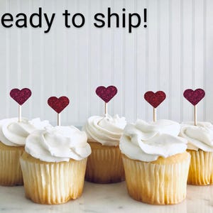 May include: Six cupcakes with white frosting and pink glitter heart toppers. The cupcakes are ready to ship.