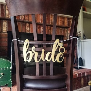 May include: A brown wooden chair with a black seat cushion. A gold glitter banner with the word "bride" and a diamond ring is strung across the back of the chair.