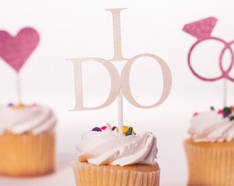 May include: Three cupcakes decorated with white frosting and colorful sprinkles. One cupcake has a pink heart topper, another has a white "I DO" topper, and the third has a pink ring topper.