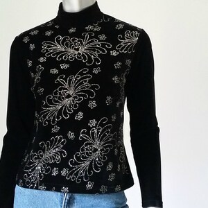 Puede incluir: Top negro de manga larga con cuello alto. El top est&aacute; adornado con un estampado floral plateado en la parte delantera. Las mangas son largas y ajustadas. El top se combina con unos vaqueros azules.