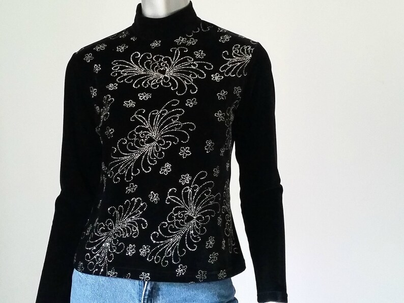 Puede incluir: Top negro de manga larga con cuello alto. El top est&aacute; adornado con un estampado floral plateado en la parte delantera. Las mangas son largas y ajustadas. El top se combina con unos vaqueros azules.