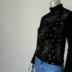 Puede incluir: Top negro de manga larga con cuello alto. La parte superior presenta un intrincado dise&ntilde;o floral plateado en la parte delantera. La modelo lleva vaqueros azules. La parte superior est&aacute; hecha de un material suave y c&oacute;modo.