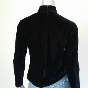 Puede incluir: Un top negro de manga larga de terciopelo con cuello alto y cremallera en la espalda. La prenda se exhibe en un maniqu&iacute;, mostrando la vista trasera. La tela es suave y tiene un ligero brillo.