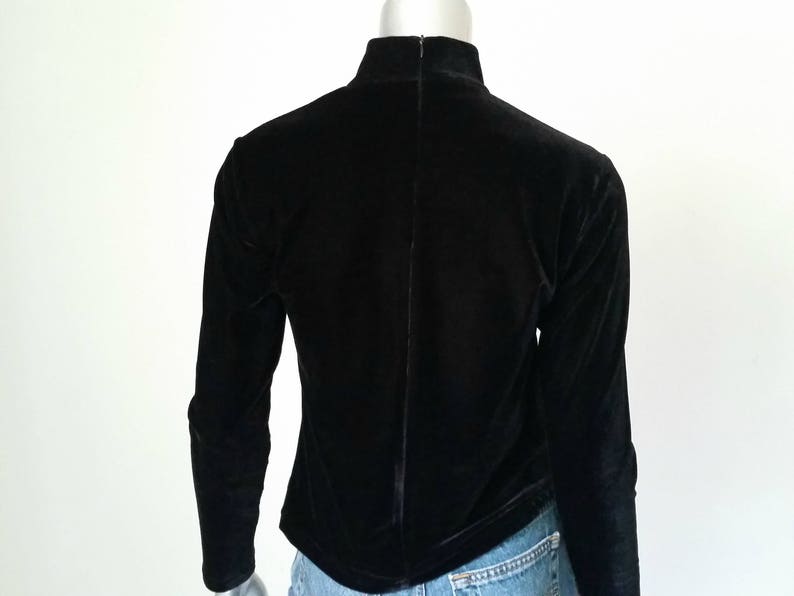 Puede incluir: Un top negro de manga larga de terciopelo con cuello alto y cremallera en la espalda. La prenda se exhibe en un maniqu&iacute;, mostrando la vista trasera. La tela es suave y tiene un ligero brillo.