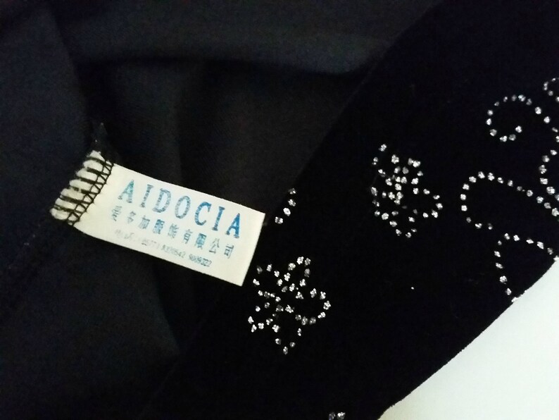 Puede incluir: Tela negra con un patr&oacute;n decorativo plateado. Una etiqueta blanca con la marca "AIDOCIA" y otro texto es visible. La tela parece ser parte de una prenda.