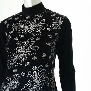 Puede incluir: Top negro de manga larga con cuello alto. El top est&aacute; adornado con un intrincado dise&ntilde;o floral plateado en el pecho y los hombros. El dise&ntilde;o est&aacute; hecho de peque&ntilde;os puntos detallados y l&iacute;neas curvas.