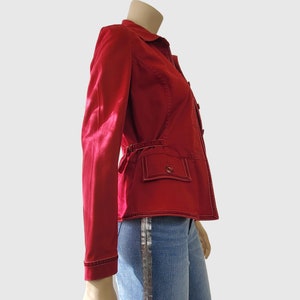 May include: A red jacket with a collared neckline, long sleeves, and a button-up front. The jacket features two front pockets with button closures and a belt detail at the waist. White stitching accents the edges. The jacket is paired with blue jeans.
