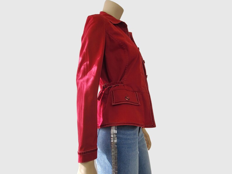 May include: A red jacket with a collared neckline, long sleeves, and a button-up front. The jacket features two front pockets with button closures and a belt detail at the waist. White stitching accents the edges. The jacket is paired with blue jeans.