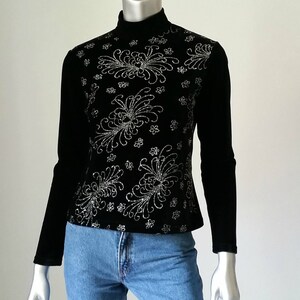 Puede incluir: Top negro de manga larga con cuello alto. La parte delantera del top est&aacute; decorada con un estampado floral plateado. El top se combina con unos vaqueros azules. El top est&aacute; hecho de un material suave, similar al terciopelo.