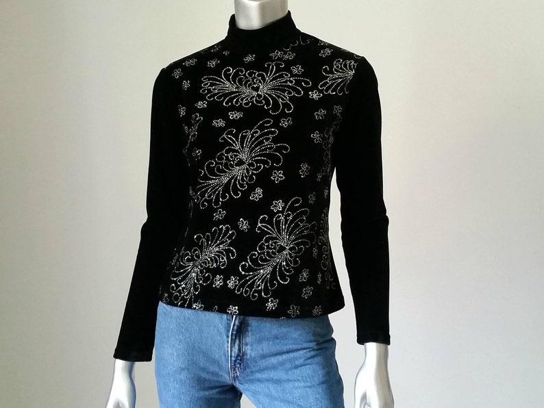 Puede incluir: Top negro de manga larga con cuello alto. La parte delantera del top est&aacute; decorada con un estampado floral plateado. El top se combina con unos vaqueros azules. El top est&aacute; hecho de un material suave, similar al terciopelo.
