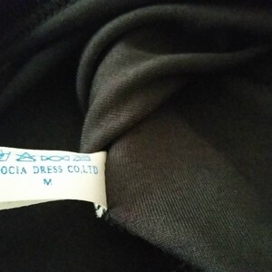 Puede incluir: Primer plano de una tela negra con una etiqueta blanca. La etiqueta tiene texto azul que dice "AIDOCIA DRESS CO.LTD" y la talla "M". La tela es oscura, posiblemente negra, y parece ser tejida.
