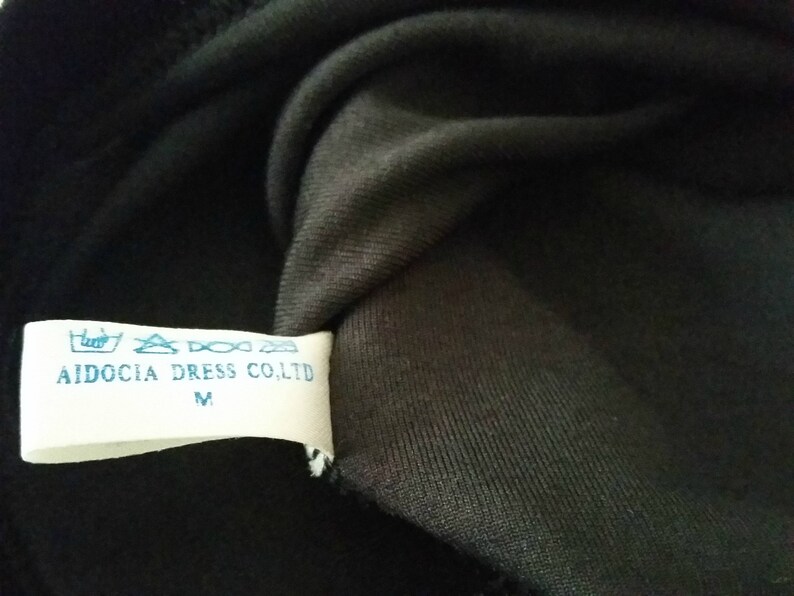 Puede incluir: Primer plano de una tela negra con una etiqueta blanca. La etiqueta tiene texto azul que dice "AIDOCIA DRESS CO.LTD" y la talla "M". La tela es oscura, posiblemente negra, y parece ser tejida.