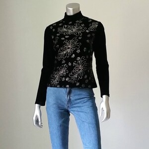 Puede incluir: Top negro de manga larga con cuello alto. El corpi&ntilde;o est&aacute; adornado con bordados florales plateados. El top se combina con vaqueros de mezclilla azul claro. El maniqu&iacute; se muestra sobre un fondo blanco liso.