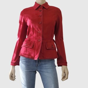 May include: A red jacket with white stitching and button fastenings. The jacket features two front pockets and a small belt detail at the waist. The model is wearing blue jeans.