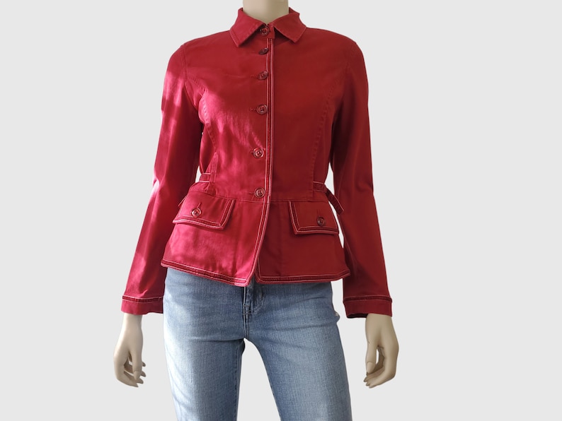 May include: A red jacket with white stitching and button fastenings. The jacket features two front pockets and a small belt detail at the waist. The model is wearing blue jeans.