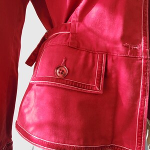 May include: A close-up of a red jacket with white stitching details. The jacket has a pocket with a button closure and a belt. The stitching outlines the pocket and edges of the jacket, creating a contrasting design element.