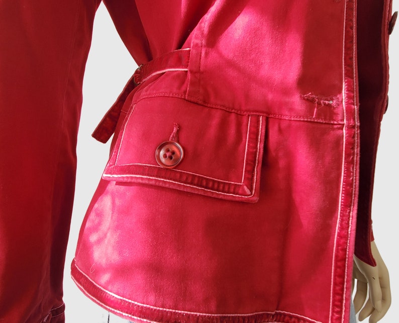 May include: A close-up of a red jacket with white stitching details. The jacket has a pocket with a button closure and a belt. The stitching outlines the pocket and edges of the jacket, creating a contrasting design element.