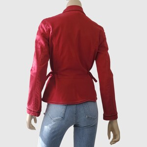May include: A red jacket with long sleeves and a wrap-around tie closure. The jacket has a collar and white stitching details. The model is wearing blue jeans. The jacket is displayed on a mannequin.
