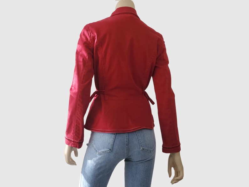 May include: A red jacket with long sleeves and a wrap-around tie closure. The jacket has a collar and white stitching details. The model is wearing blue jeans. The jacket is displayed on a mannequin.