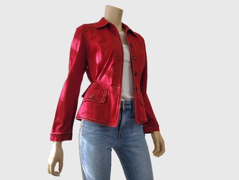 May include: A red jacket with a button-up front, two front pockets, and white stitching. The jacket is paired with blue jeans. The jacket has a collar and long sleeves. The model is wearing a white shirt underneath the jacket.