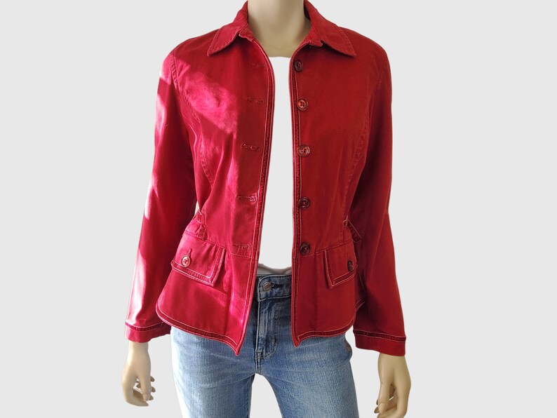May include: A red jacket with a button-up front, collar, and two front pockets. The jacket has white stitching details and is worn over a white top and blue jeans. The jacket is displayed on a mannequin.