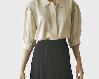 May include: Cream-colored blouse with a subtle pattern, featuring a collared neckline and three-quarter sleeves. The blouse is paired with a black skirt. The sleeves have a slight puff detail at the shoulders.