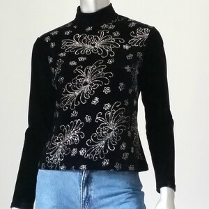 Puede incluir: Top negro de manga larga con cuello alto. El top presenta un intrincado patr&oacute;n floral plateado en el pecho y los hombros. Las mangas son negras lisas, combinado con unos vaqueros azules.