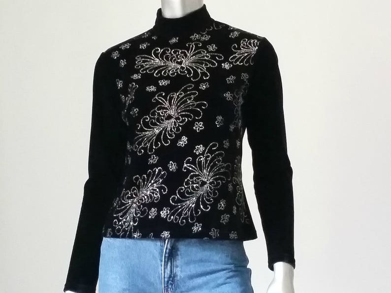 Puede incluir: Top negro de manga larga con cuello alto. El top presenta un intrincado patr&oacute;n floral plateado en el pecho y los hombros. Las mangas son negras lisas, combinado con unos vaqueros azules.