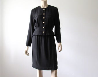 May include: A black two-piece suit displayed on a mannequin. The jacket has long sleeves, a round neckline, and gold-colored buttons. The skirt is knee-length. The suit is a classic style, suitable for formal or professional settings.