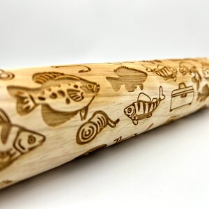May include: A wooden rolling pin with a fishing theme. The rolling pin is engraved with images of fish, fishing lures, and a tackle box.