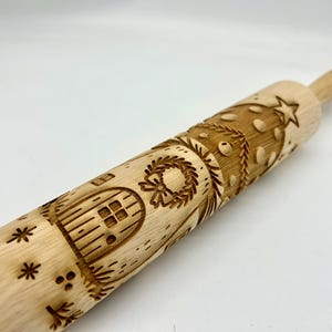 May include: A wooden rolling pin with engraved Christmas-themed designs. The pin features a Christmas tree, a house with a door, a wreath, and snowflakes. The rolling pin is made of light-colored wood.
