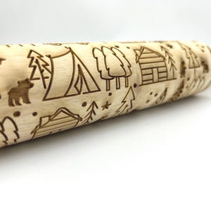 May include: A wooden rolling pin with an engraved design of a camping scene. The design includes trees, a tent, a cabin, and a bear.