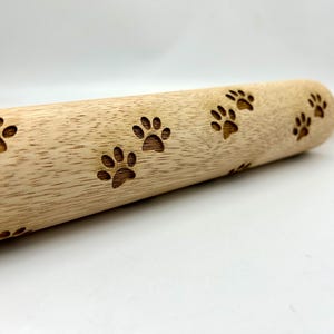 May include: A wooden rolling pin with a paw print design. The rolling pin is light brown and has a smooth surface. The paw prints are evenly spaced and are a darker brown color.