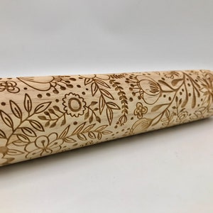 May include: A light-colored wooden rolling pin with a floral design carved into its surface. The design includes various flowers, leaves, and dots. The rolling pin has a smooth, light-colored wooden handle on each end.