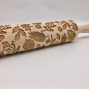 May include: A wooden rolling pin with an engraved design of leaves, acorns, and branches. The rolling pin is light brown and has a natural wood finish.