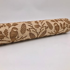 May include: A wooden rolling pin with an engraved design of birds, holly leaves, and pine cones. The design is in a brown color and is repeated across the entire surface of the rolling pin.