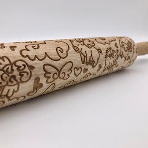May include: A wooden rolling pin with a carved design of pigs with wings and hearts. The rolling pin is made of natural wood and has a light brown color.