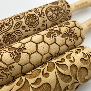 May include: Three wooden rolling pins with different engraved designs. The top rolling pin has a paisley pattern, the middle rolling pin has a honeycomb pattern with bees, and the bottom rolling pin has a floral pattern.
