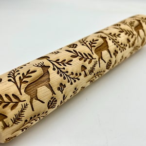 May include: A wooden rolling pin with a detailed engraved design. The design features deer, leaves, and mushrooms. The rolling pin is light brown and has a smooth, polished finish. The handles are a lighter shade of wood.
